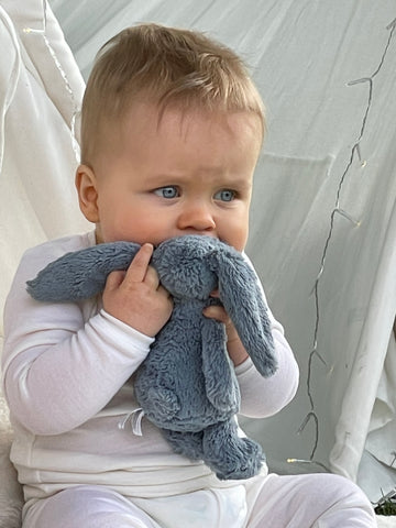 Baby in merino wool pyjamas, sits holding a blue cuddly rabbit toy up to their mouth.