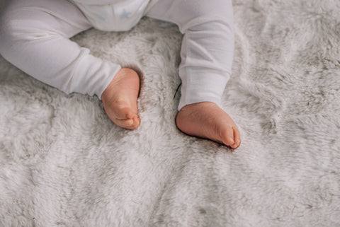 A bird's eye view of a sitting baby's legs wearing ivory pyjamas on a white blanket.