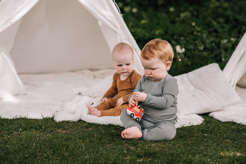 Two babies sit playing, outside a tent on the grass, wearing merino pyjamas.