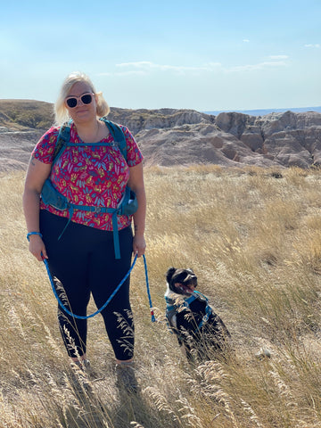 Jenny (founder of Unlikely Hikers) and Big Judy (Border Collie mix) take a break while hiking in a grassy field with rocks behind.