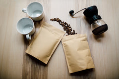 Coffee grinder and two packages of coffee beans with two white mugs on table