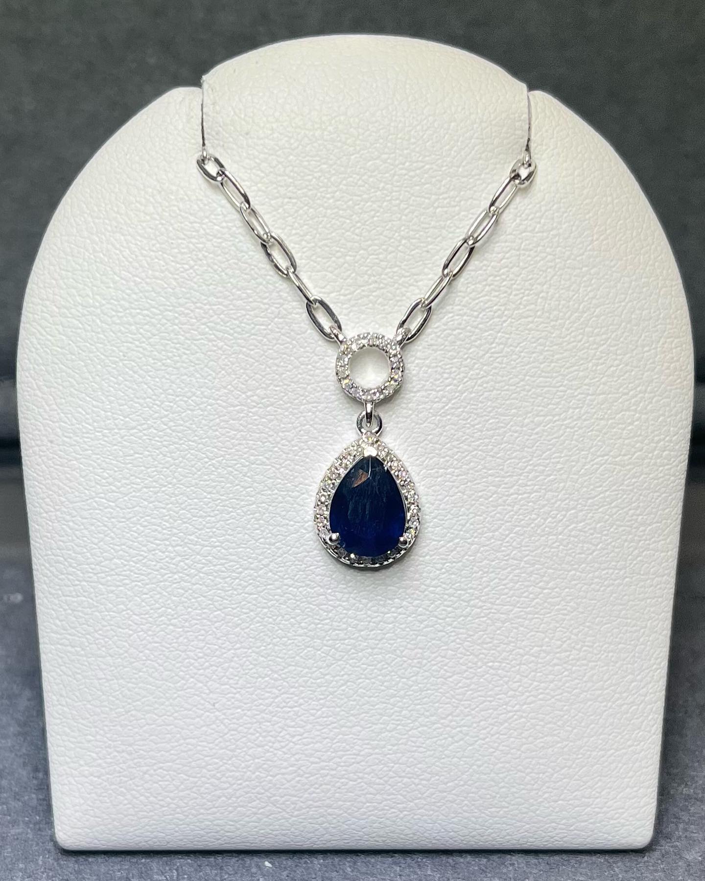Silver necklace with a pear shaped blue gemstone display on a white jewelry holder