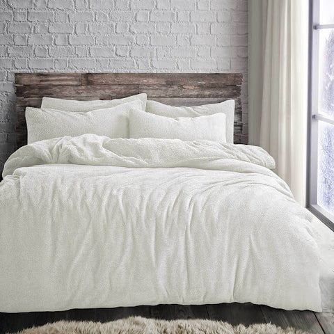 A cosy and inviting bed set in a modern bedroom featuring a plush, cream-coloured duvet from Shaws, complemented by matching pillows. The bed is set against a rustic wooden headboard and a white brick wall, with natural light streaming in from a window to the side.