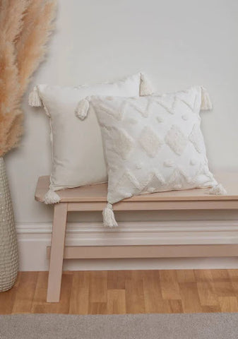 wo white cushions on top of a bench. Both cushions have tassels in the corners, and one has diamond detailing.