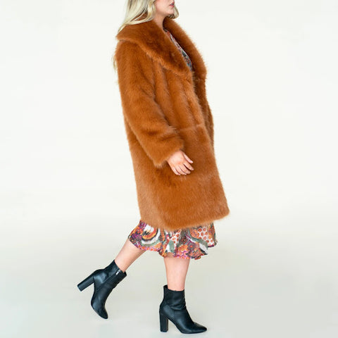 The model wears a tan faux-fur coat over a brightly coloured dress with black leather ankle boots
