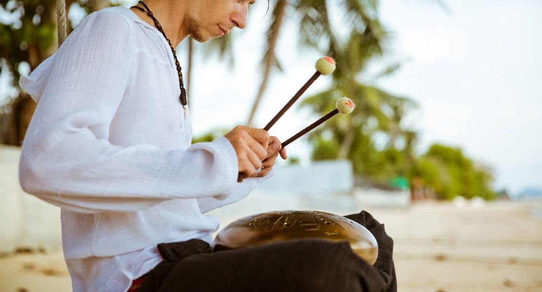 Un homme assis sur la plage jouant du Tongue Drum avec des mailloches
