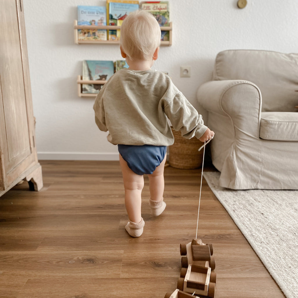 Bébé Judes marchant avec un jouet à tirer près d'une étagère avec des livres en maternelle