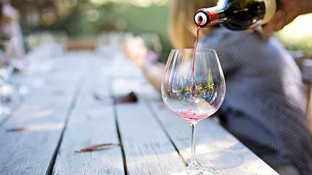 Hand pouring red wine into a tall, rounded wine glass.