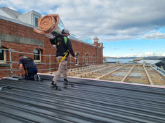 This is a photo of two workers on a roof installing new Stramit Speed Deck Ultra Roof materials supplied by Roofing Supplies Online on a commercial job site. They are wearing Safety Harnesses & there is also a safety rail around the roof perimeter.