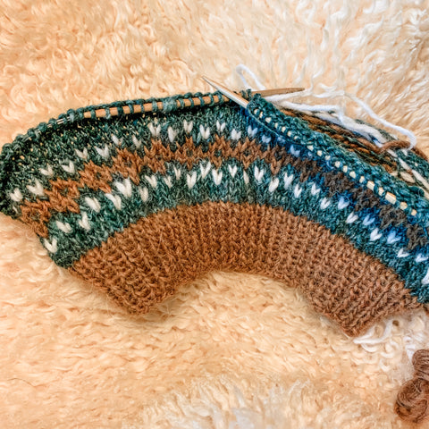 Bottom of sweater on bamboo needles lamb's fleece background. Yarn colors are chestnut, teal, and white. There is a colorwork pattern.