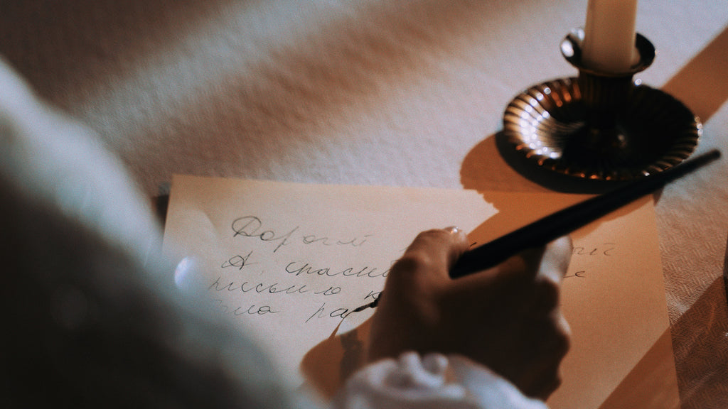 A Green Witch in a billowy white blouse journals with a quill pen in a dark moody candlelit room.