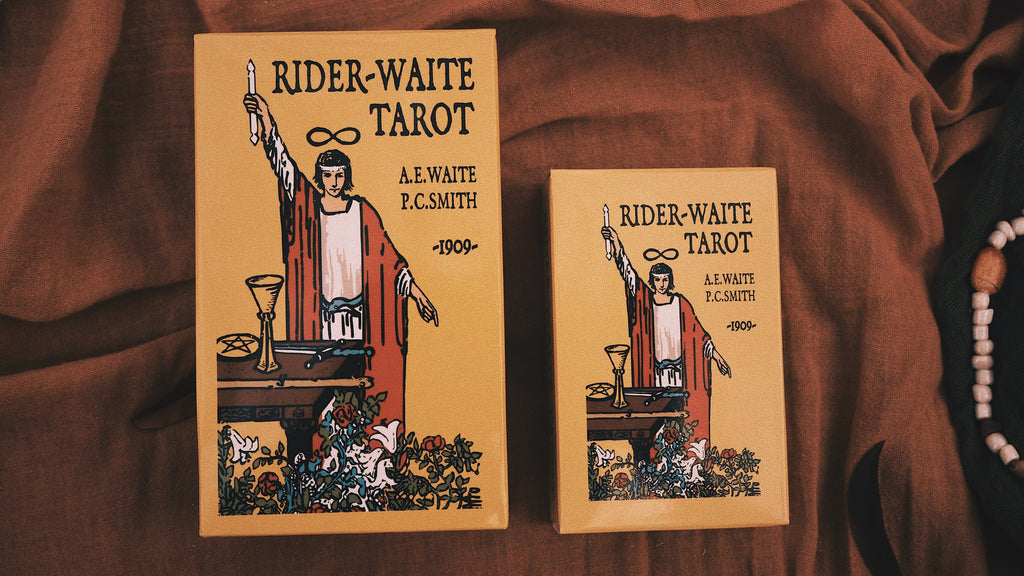 wo editions of the Rider-Waite Tarot deck rest against a backdrop of warm-toned fabric. The tarot covers feature a symbolic illustration of a figure with raised hand holding a candle, surrounded by spiritual elements like a chalice and pentacle. To the side, a beaded strand, reminiscent of runes, lies partially visible. These mystical tools suggest a deeper exploration and guidance during the powerful Full Blood Moon in Taurus phase