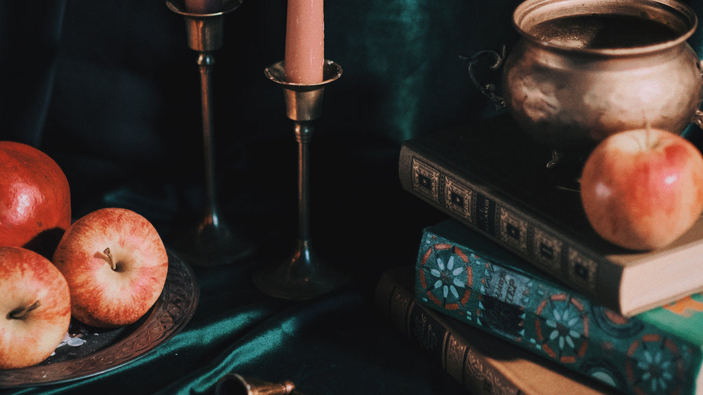 A scene of red/gold apples, peachy colored candles, and books against a dark green velvety backdrop.