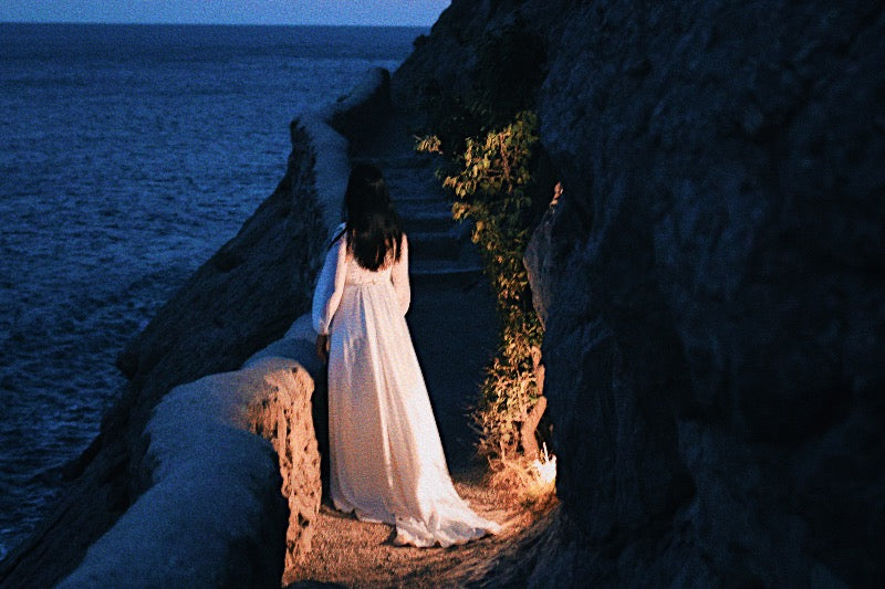 A woman stands in a white flowy cottage core dress on a dark seaside cliff overlooking the ocean.