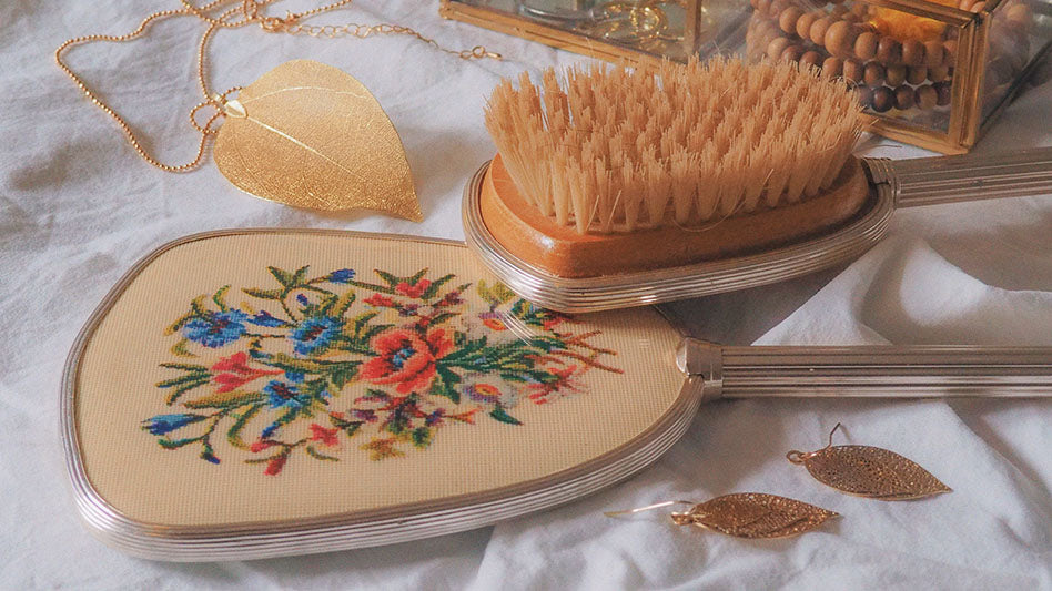 Collection of Aphrodite's symbols including jewelry, embroidered floral mirror, wooden brush, and beaded earrings on a delicate fabric backdrop