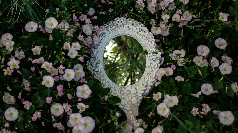 A decorative round mirror with a detailed white frame, reflecting a lush backdrop of light pink morning glory flowers, symbolizing self-reflection and the blossoming possibilities of the New Moon in Aries.