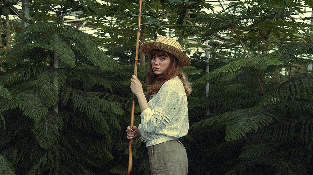 A woman in a vintage outfit stands contemplatively in a lush fern greenhouse, resonating with the introspective energy of the new moon.