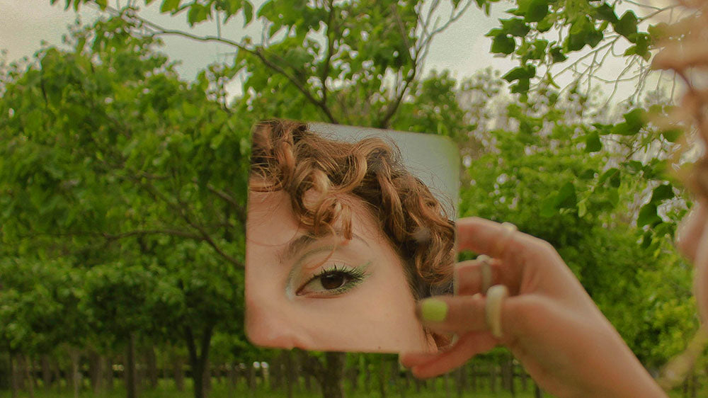 A person holds a small mirror reflecting their eye and curls, amidst a verdant garden scene, suggesting self-reflection and the personal insights available during the Aries New Moon.