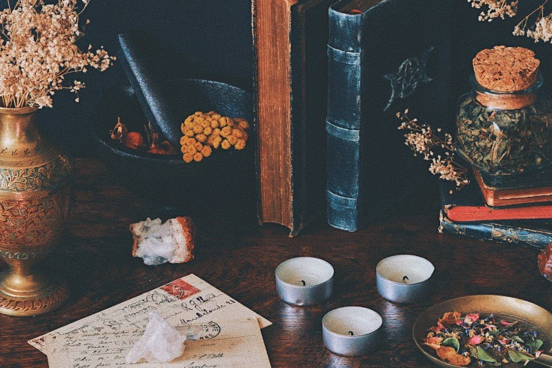 A moody still life depicting the history of chaos magic, featuring old books on esoteric knowledge, a mortar and pestle for herbal preparations, flickering candles, magical instruments, and dried flowers, all artfully arranged on a vintage wooden table.
