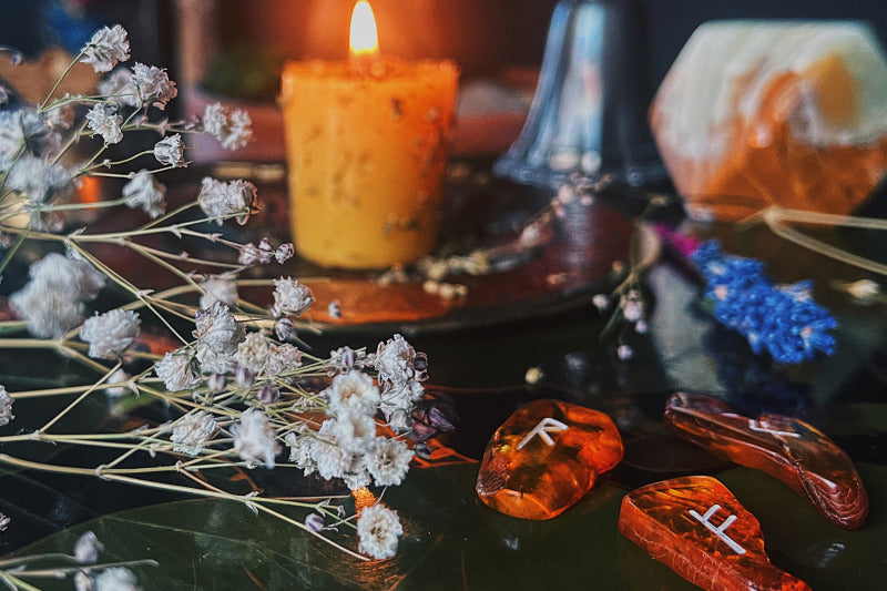 A mystical array of amber runes for chaos magic divination spread out on a asian lacquer table, illuminated by the inviting glow of a single candle. Delicate sprigs of baby's breath flowers add an enchanting touch to this earth witch practice scene