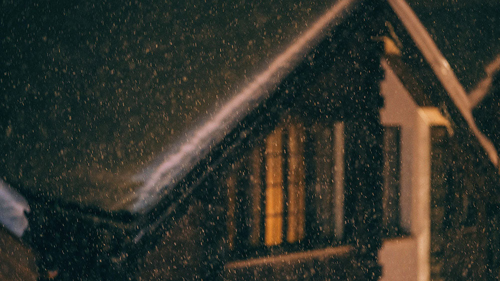 A night scene showing heavy snowfall with a warm yellow light glowing from the windows of a dark house. The snowflakes are in sharp focus against a blurry backdrop, with a part of the house's roof and a streetlight visible in the frame.