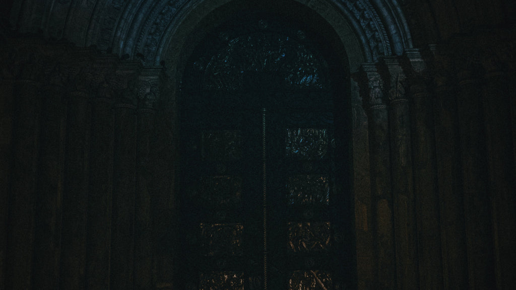 A dimly lit image of an ornate, heavy-set door flanked by sculpted columns, suggestive of an entrance to a temple or ancient site. This evocative scene represents the threshold of the chthonic realm, a domain often associated with Hekate, the goddess who stands at the portals between worlds.