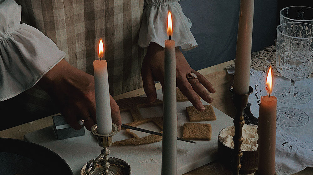 A dimly lit scene with hands working on a kitchen table during ritual preparation, featuring lit white candles, shortbread cookies, and crystal glassware, invoking a sense of calm and tradition.