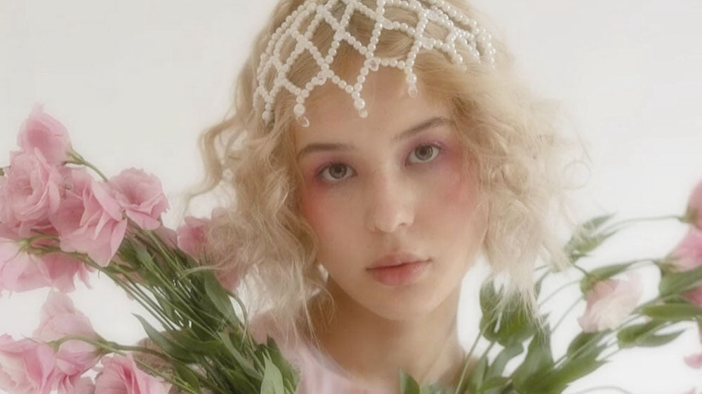A dreamy portrait of a young woman adorned with a pearl headpiece and surrounded by soft pink flowers, her ethereal gaze and the floral ambiance suggesting a connection to divine femininity and nature spirits.