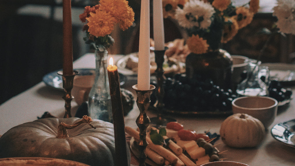 Image showcases an atmospheric and intimate table setting, evoking the essence of the Full Blood Moon in Taurus and Samhain celebrations. At the forefront, a pale gray pumpkin rests, its twisted stem curling upwards, suggesting the season's harvest. Clustered on the table are vintage candlesticks of varying heights, one holding a burnt-out candle and another with a tall, glowing white one, casting a soft luminescence. Amidst the dim light, richly colored bouquets of marigold and white flowers rise in rustic glass jars, adding vibrancy and warmth. Scattered around are an assortment of foods: a plate of dark grapes, freshly baked bread rolls, and a wooden skewer threaded with cheese and cherry tomatoes. The ambience suggests a ritual or feast, capturing the deep connection to nature and ancestors during this spiritual time.