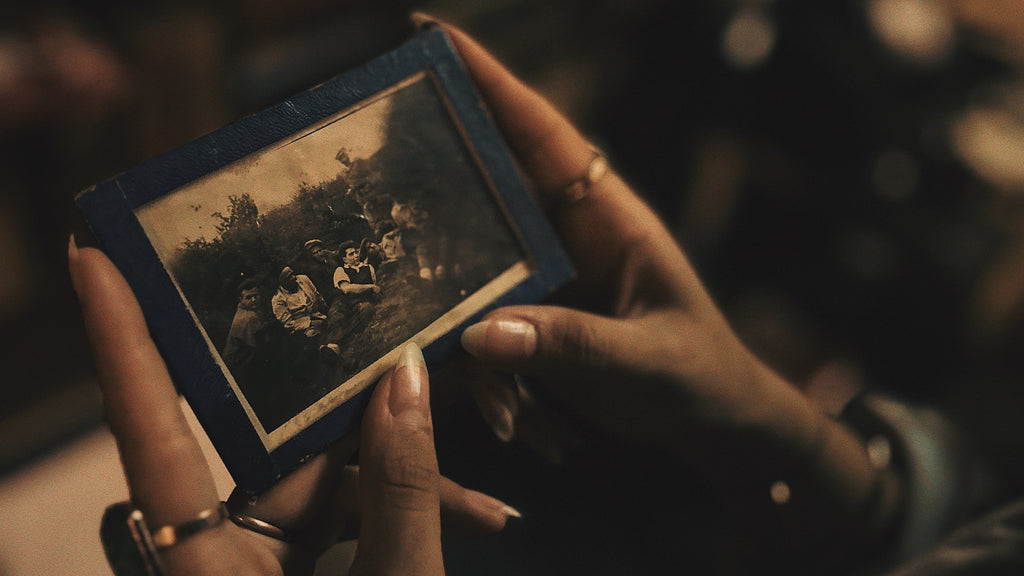 Image captures a pair of gently-held hands cradling an old, faded photograph framed in a weathered blue border. The photograph reveals a group of individuals, presumably ancestors, seated in a pastoral setting, their faces telling tales of days long past. The surrounding ambiance is dimly lit, creating a sense of intimacy and reverence. The delicate rings on the holder's fingers and the soft focus in the background amplify the mood of remembrance, emphasizing the act of connecting with one's lineage during the powerful energy of the Full Blood Moon in Taurus.