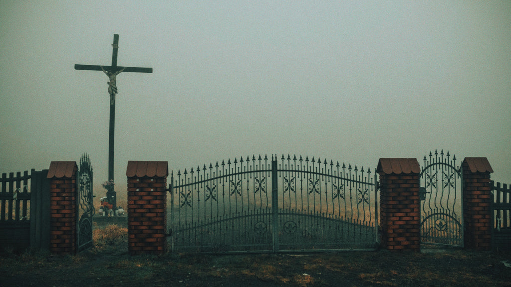 An overcast view of a wrought-iron gate leading to a cemetery, with a large cross towering in the background. The misty atmosphere and somber setting evoke Hekate's association with the threshold between the worlds of the living and the dead.