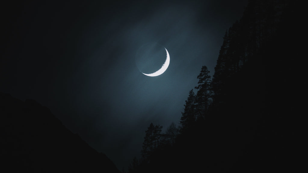 A crescent moon casting a silvery glow through a night sky, with the silhouette of a forested mountain landscape below. This image evokes the mystical association of the crescent moon with Hekate, the goddess of magic and the night, highlighting her connection to the lunar cycle and the enigmatic power of the natural world.