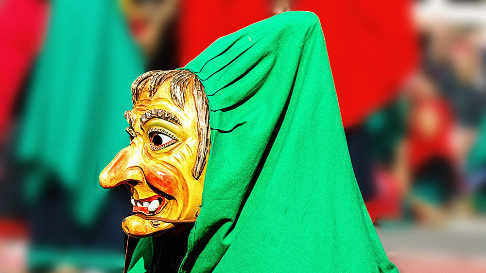 A vibrant photograph capturing a person wearing a traditional wooden mask with exaggerated features, covered by a bright green cloak. The mask portrays a comical or possibly grotesque expression, with the scene likely part of a festival or cultural event.