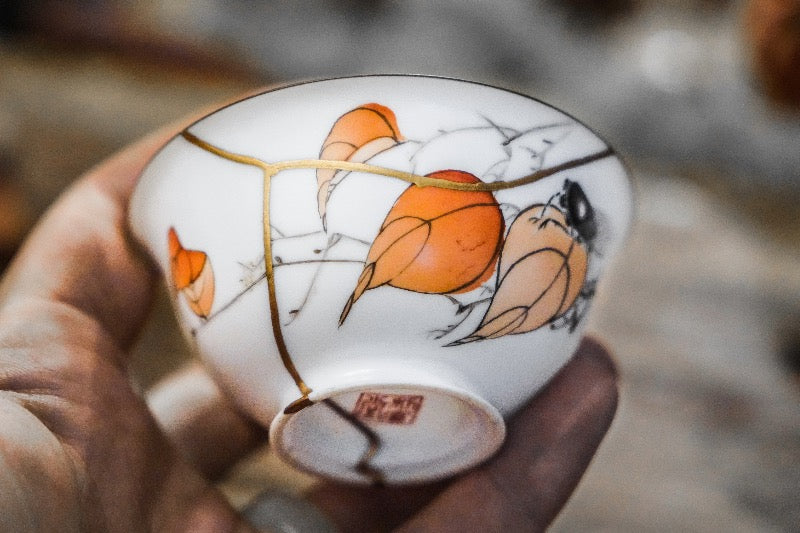 Close-up of a hand holding a delicate white porcelain teacup, adorned with orange leaves and fine gold kintsugi repair lines, symbolizing the beauty and stories behind cherished objects.