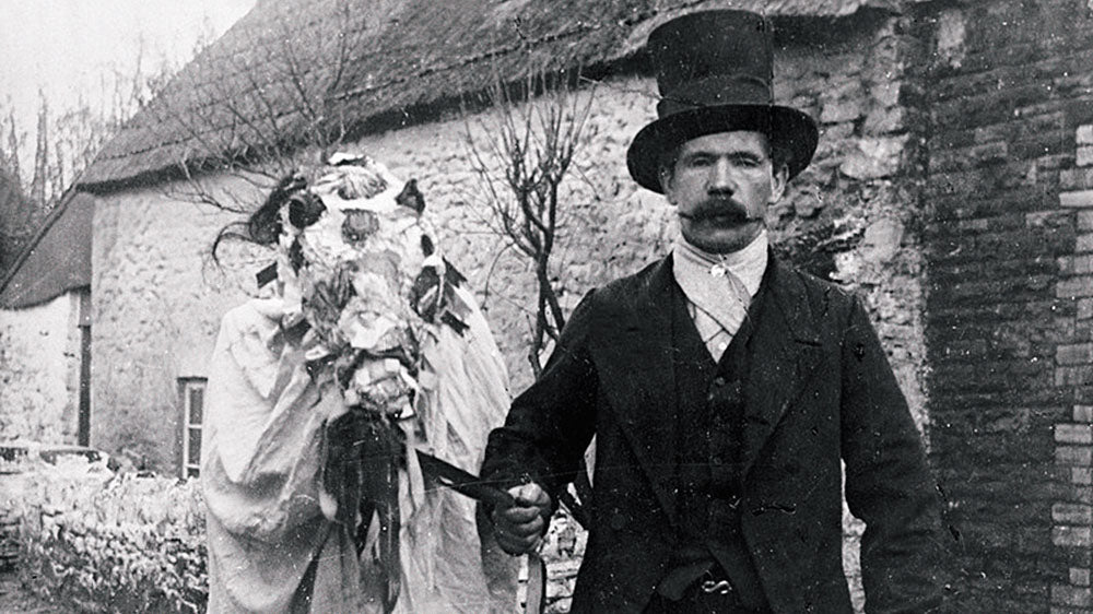 This is a black and white photograph depicting two figures in a rural setting. On the left, there is a person obscured by a large, ornate horse skull decorated with ribbons and foliage, a part of the Welsh folk tradition of Mari Lwyd. The figure is draped in a white sheet or cloak, holding the horse skull on a stick. To the right stands a man in a dark suit, waistcoat, and tie, wearing a top hat, presenting a stern expression. The backdrop includes a stone wall and a building with a thatched roof, suggesting this photo might have been taken in the early 20th century or late 19th century, during a winter festival or folk celebration.
