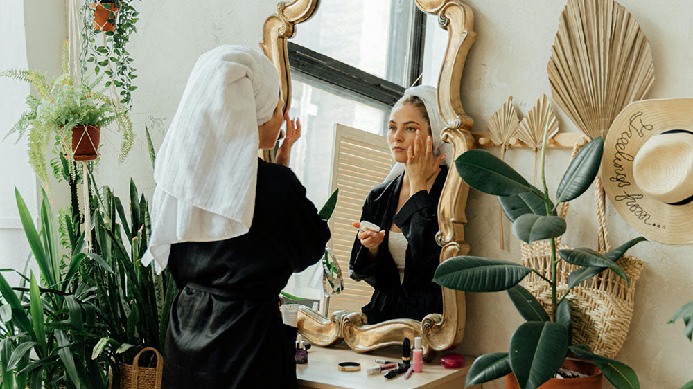 A woman in a bathroom mirror applying skincare, surrounded by lush green plants, reflecting self-care and the balance of inner and outer beauty.