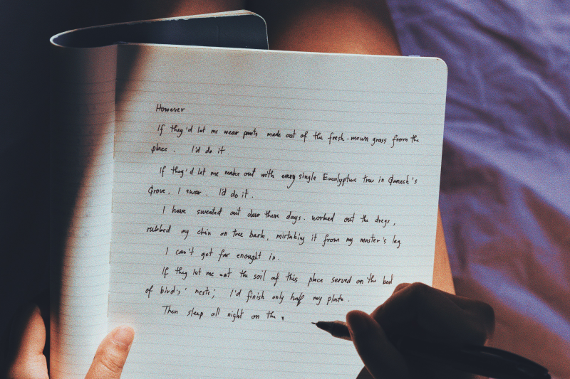 A woman journals her thoughts onto a notebook while sitting comfortably on a purple bed.