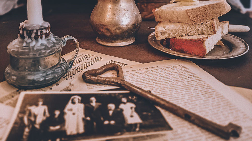 A vintage-styled photograph featuring an old candle holder, a stack of sandwiches, and black-and-white family pictures spread out on a table with a vintage book and a musical instrument, evoking a sense of ancestral connection during the New Wolf Moon.