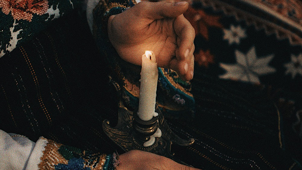 A close-up of a woman's hands gently shielding a flickering candle flame, the warmth of the light contrasting with the rich, dark textiles of her traditional clothing. This image conveys the contemplative energy of the New Oak December Moon in Sagittarius, symbolizing the light of wisdom and guidance during the introspective lunar cycle, and the protection of our inner flame against the cold, expansive winter night.