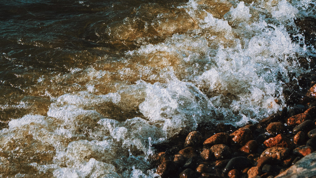 Close-up photograph of vigorous waves crashing onto a pebbled shore, symbolizing the dynamic energy of the New Oak December Moon in Sagittarius. The restless movement of the water reflects the mutable quality of Sagittarius, while the clarity of the image captures the tension of the moon squared with Neptune, suggesting a search for balance between exploration and introspection.