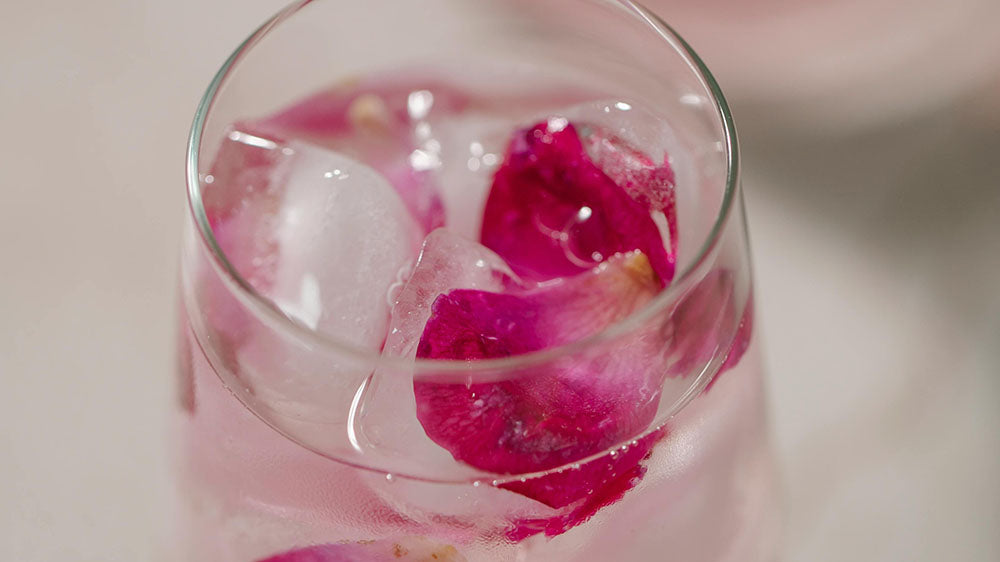Close-up of a refreshing pink beverage in a clear glass with ice cubes and vivid pink rose petals, evoking a sense of healing and heart-opening during the Pink Moon period.