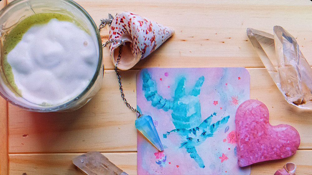 A serene and inviting tabletop scene featuring a soothing matcha latte with frothy milk, a conch shell, a pendulum with a blue crystal, a large quartz point, and a rose quartz heart. A notepad with a watercolor blue cat adds a whimsical touch.