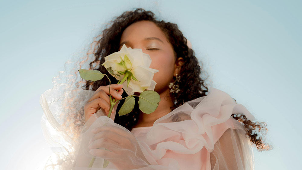 A woman in a sheer, ruffled garment inhales the fragrance of a white rose against a clear sky, embodying a moment of serene beauty and connection with nature.