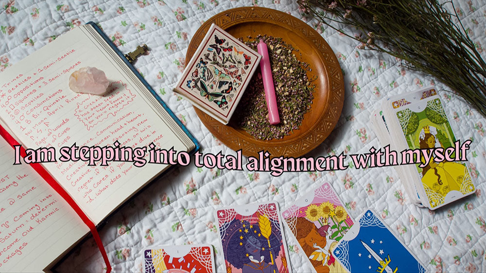 A spiritual setup featuring a tarot card spread, an open journal with red writing, dried herbs in a wooden bowl, and a pink candle, suggesting a ritual of self-discovery and alignment.