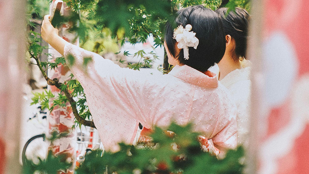 Two people in traditional yukata with their backs to the viewer, one taking a selfie with a smartphone, framed by lush greenery, capturing a modern moment in a setting of timeless tradition.