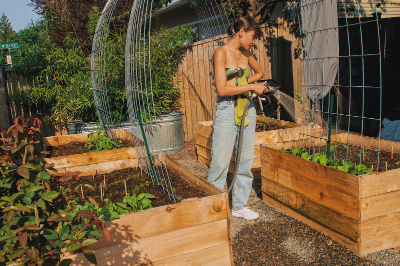 Young woman in silk scarf top and Levis, watering her vegetable garden, representing the magic blend of gardening and personal growth. Amid the radishes, carrots, kale, edible flowers, and herbs, she's planting seeds of dreams, embodying resilience, growth, and the powerful spell of manifestation.