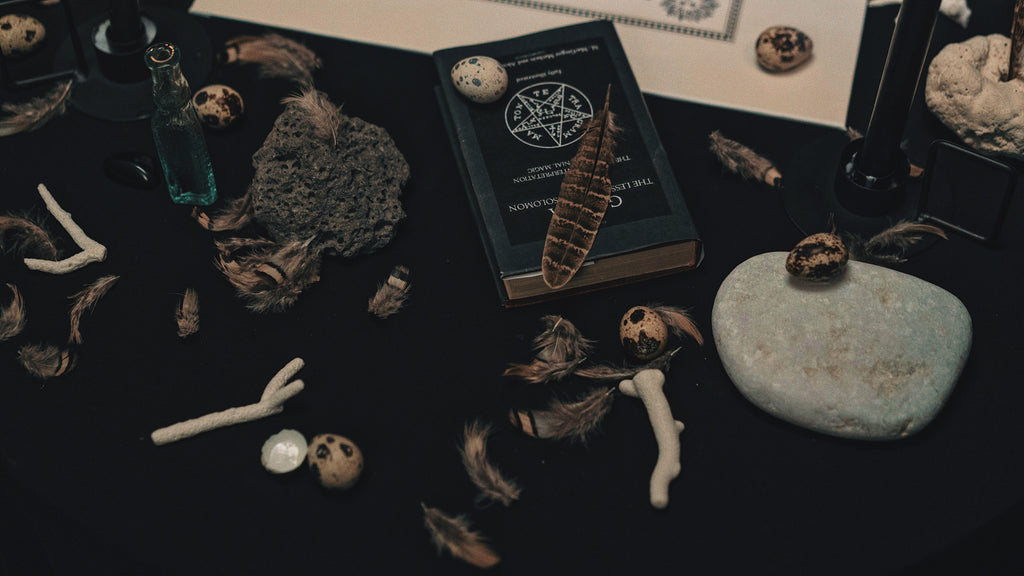 A carefully arranged altar with various symbolic objects related to Hekate. It includes feathers, quail eggs, bones, a stone slab, an antique book with a pentagram, and bottles of dark liquids. This collection creates a sacred space that resonates with the themes of witchcraft and the mystical practices associated with the goddess Hekate.