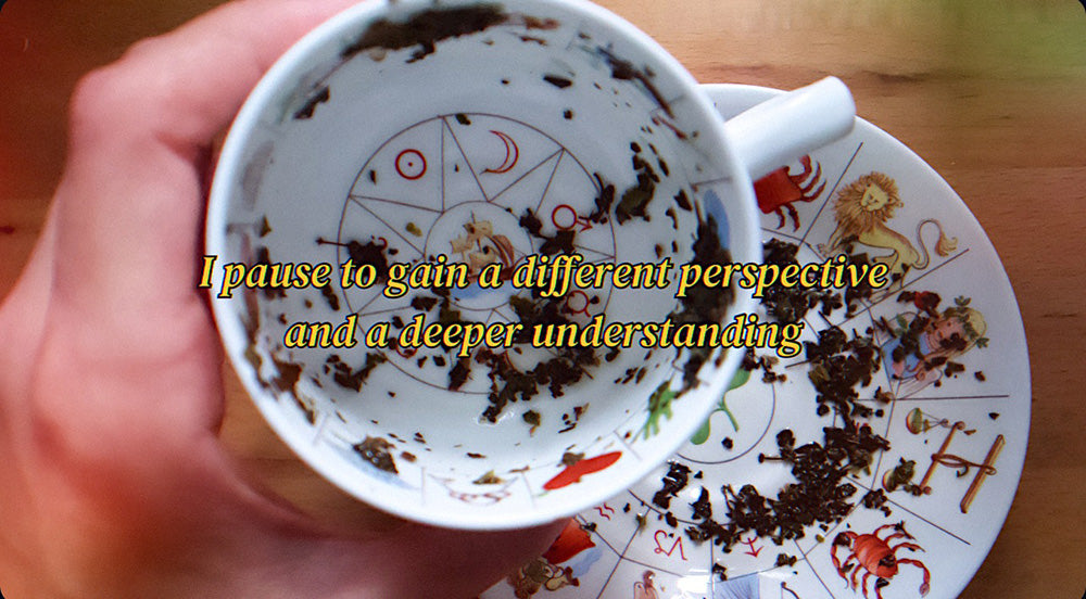 A hand holding a teacup with tea leaves settled at the bottom, above a saucer with zodiac signs and planetary symbols, with the text "I pause to gain a different perspective and a deeper understanding" overlaying the image.