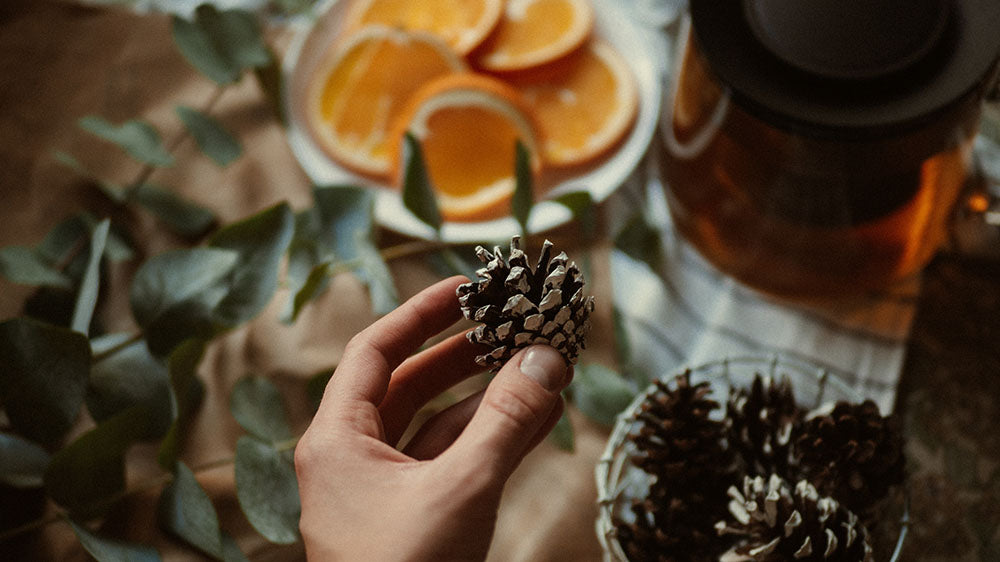 A hand holds a pinecone above a Yule altar setting, with slices of dried orange on a plate, eucalyptus leaves scattered around, and a pot of tea with a black lid in the background, all laid out on a table with a natural beige cloth.