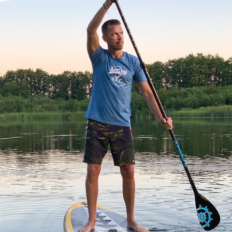 Ein junger Mann steht auf seinem SUP Board, schaut verträumt auf den See und trägt ein blaues Zurfday T-Shirt